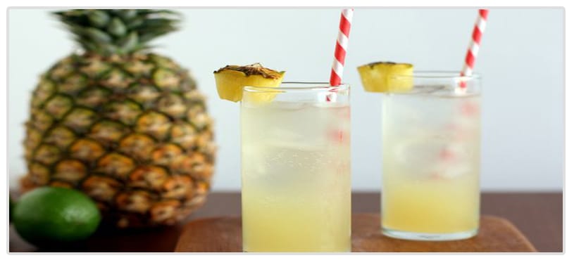 Two tall glasses of pineapple cooler with red and white striped straws sit on a wooden surface, accompanied by a fresh pineapple and a lime in the background.