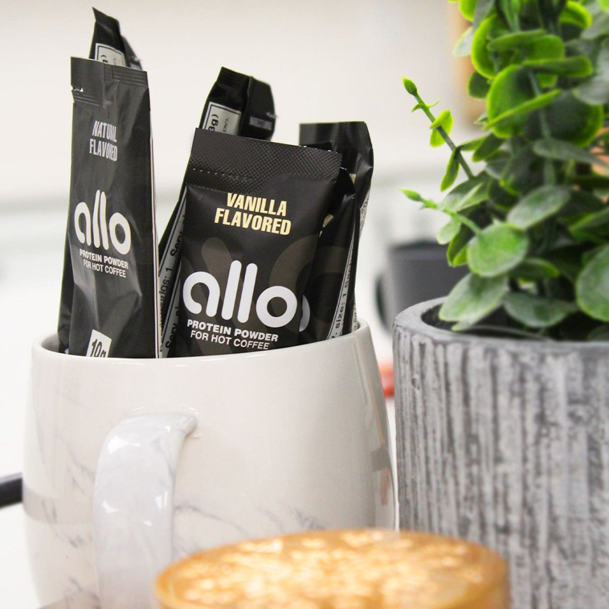 Black packets of Allo protein powder, labeled "Natural" and "Vanilla Flavored," are displayed in a white marble mug next to a textured gray planter and a blurred coffee cup in a bright setting.