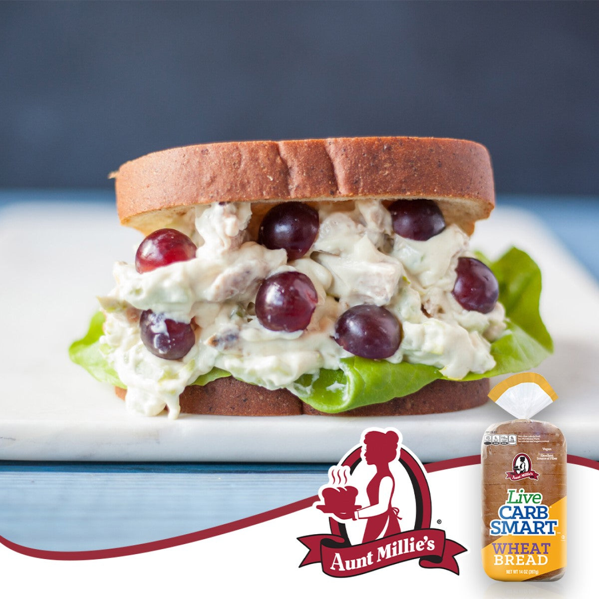 A sandwich made with Aunt Millie's Live Carb Smart wheat bread is filled with lettuce, creamy chicken salad, and grapes, set on a light blue wooden surface.