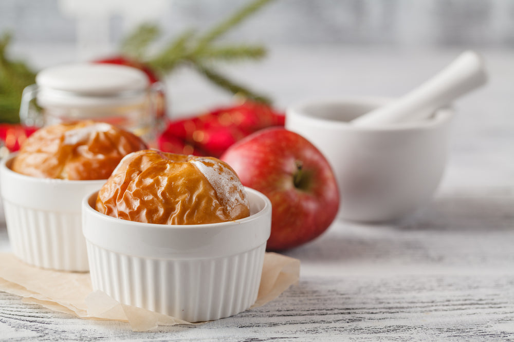 Two baked apples in white ramekins sit on a light wooden surface, with a fresh red apple and a mortar and pestle nearby, surrounded by festive decorations in the background.