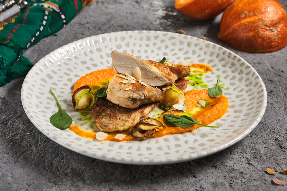 A plate features baked chicken breast sliced and garnished with slivered almonds, served alongside steamed garlic herb butternut squash puree and sautéed vegetables, with decorative green sauce and a rustic background.