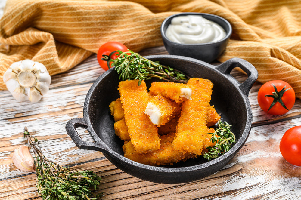 A cast iron bowl holds golden-brown baked fish sticks, garnished with fresh thyme, alongside cherry tomatoes, garlic cloves, and a small bowl of creamy dipping sauce, all set on a rustic wooden surface.