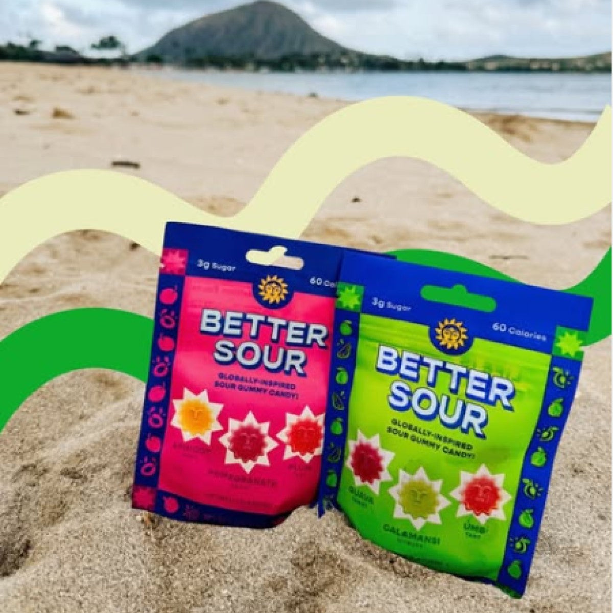 Two colorful bags of Better Sour gummy candy, one pink and one green, rest on sandy beach terrain with a scenic view of a mountain and ocean in the background, highlighting their sweet and tangy flavors.