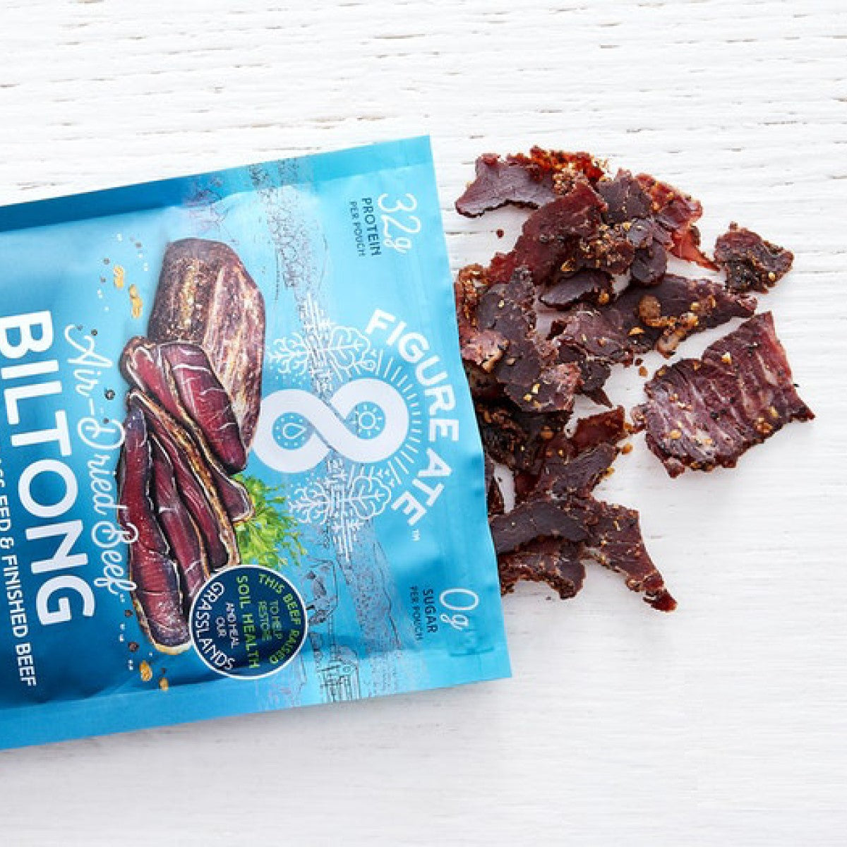 A blue package of Figure Ate air-dried beef biltong sits on a white surface, with pieces of the dried meat scattered around it, highlighting its high protein content and zero sugar.