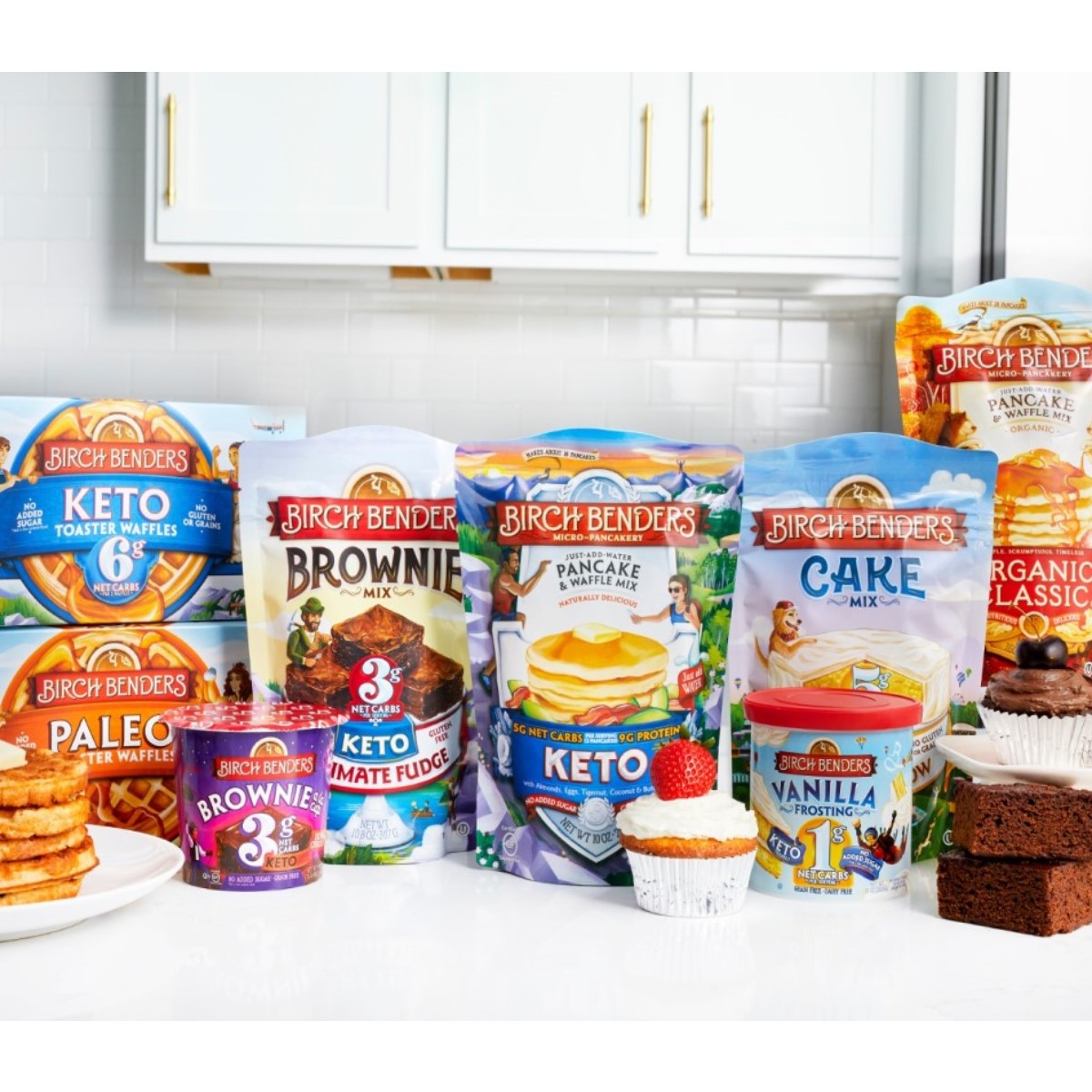 A variety of Birch Benders baking mixes and products, including pancake, brownie, and cake mixes, are displayed on a kitchen counter with finished baked goods, showcasing their branding and flavors.