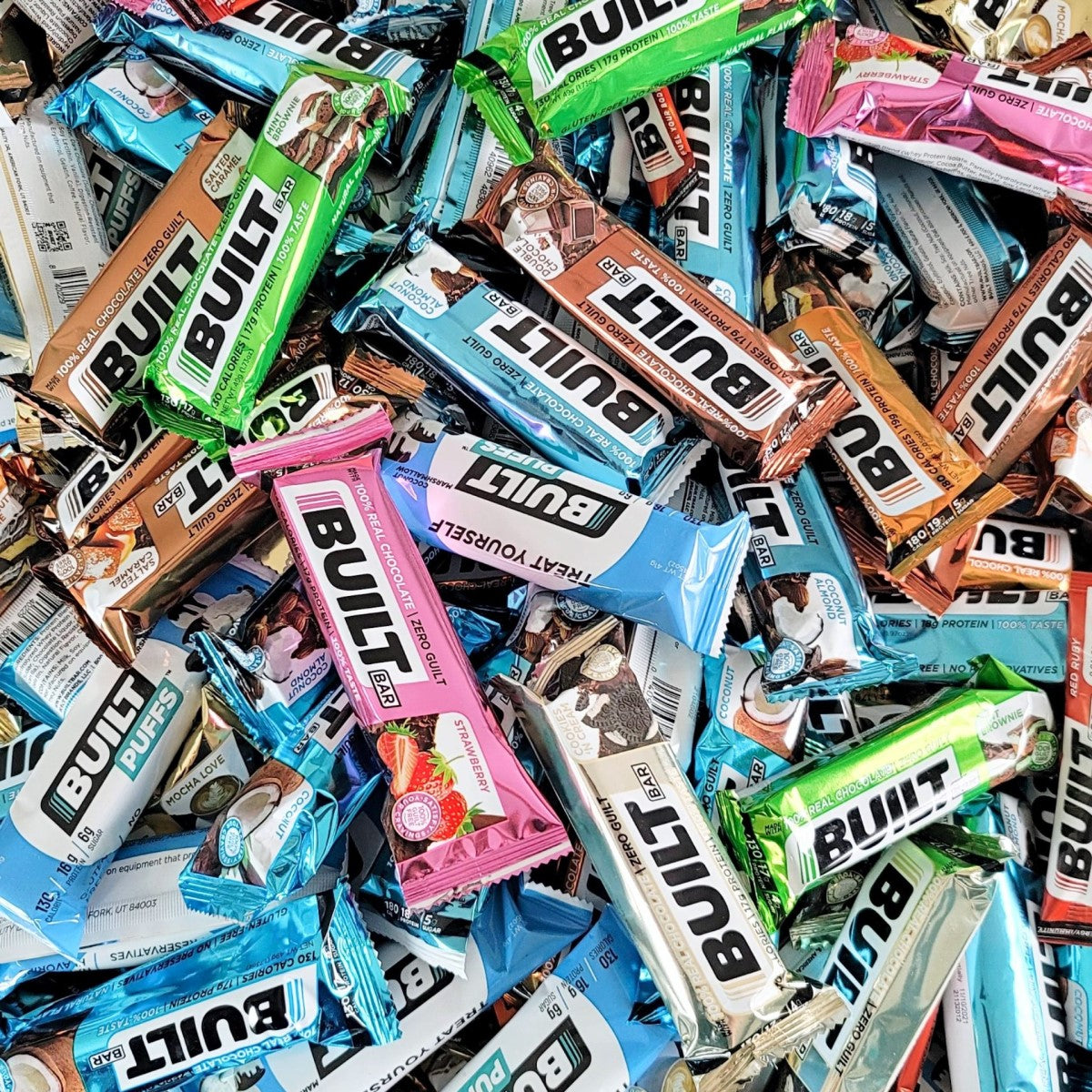 A colorful assortment of Built Bar protein bars in various flavors is scattered across a surface, showcasing their vibrant packaging and highlighting new product offerings.