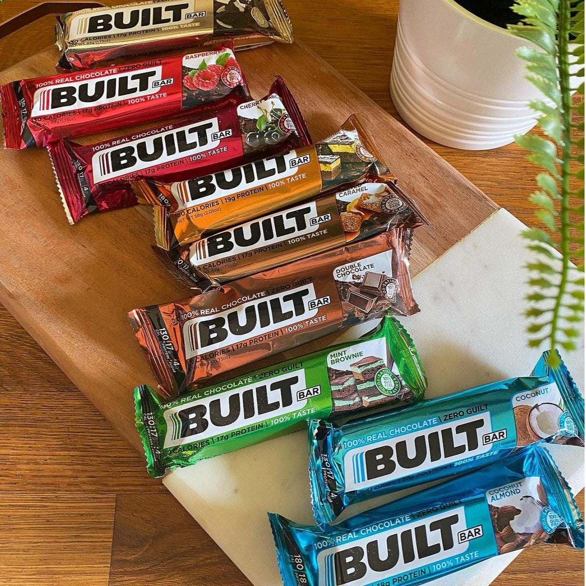 A variety of Built Bars in different flavors are neatly arranged on a wooden cutting board, with a small potted plant visible in the background, showcasing popular protein snack options.