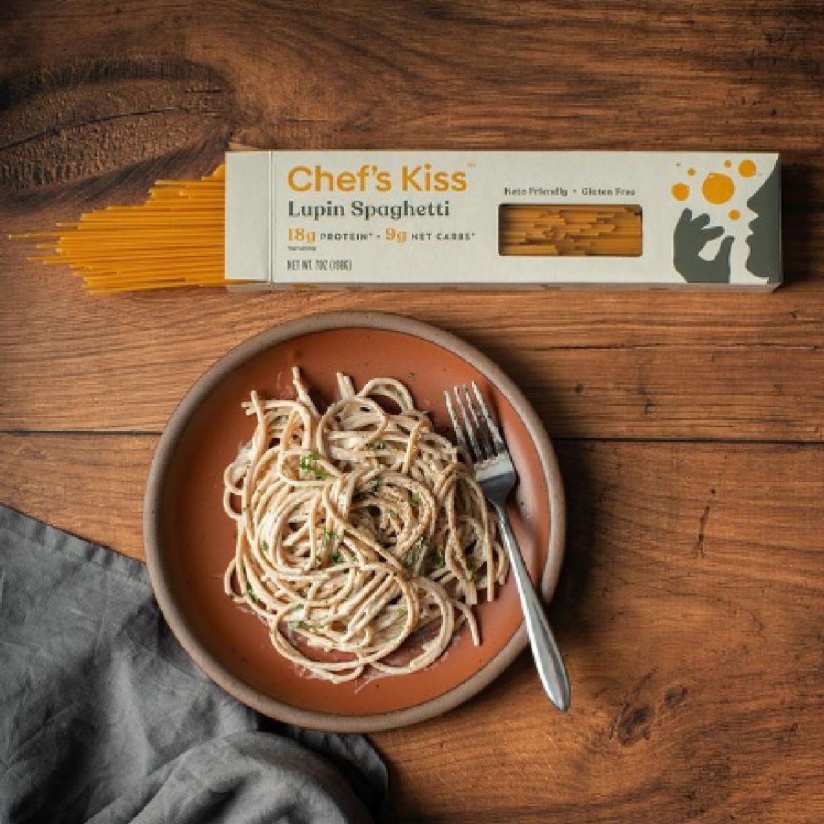 A plate of cooked lupin spaghetti topped with herbs sits next to a box labeled "Chef's Kiss" on a wooden table, with a fork resting on the plate and a gray cloth nearby.