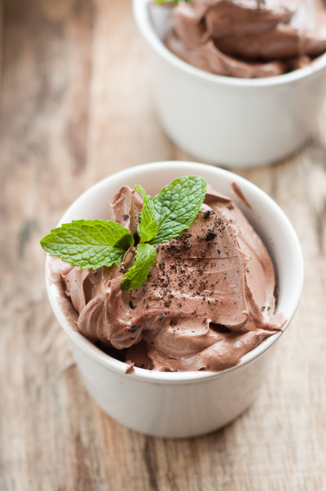 A creamy chocolate mint mousse is served in a white bowl, topped with fresh mint leaves and chocolate shavings, resting on a rustic wooden surface.