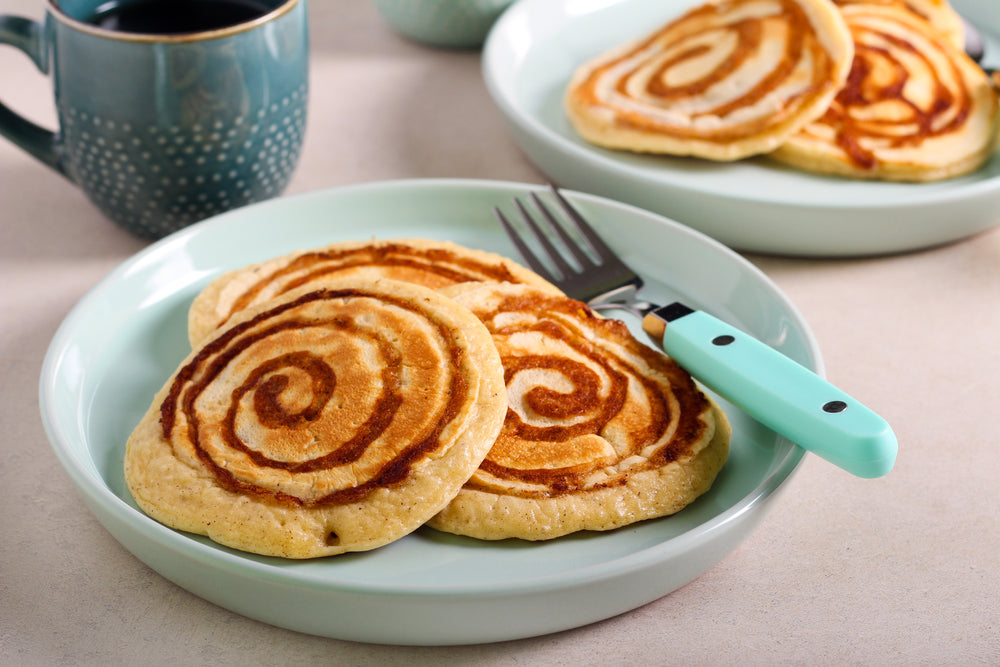 Two cinnamon bun protein pancakes with spiral patterns are served on a light green plate alongside a fork and a cup of coffee in a cozy kitchen setting.
