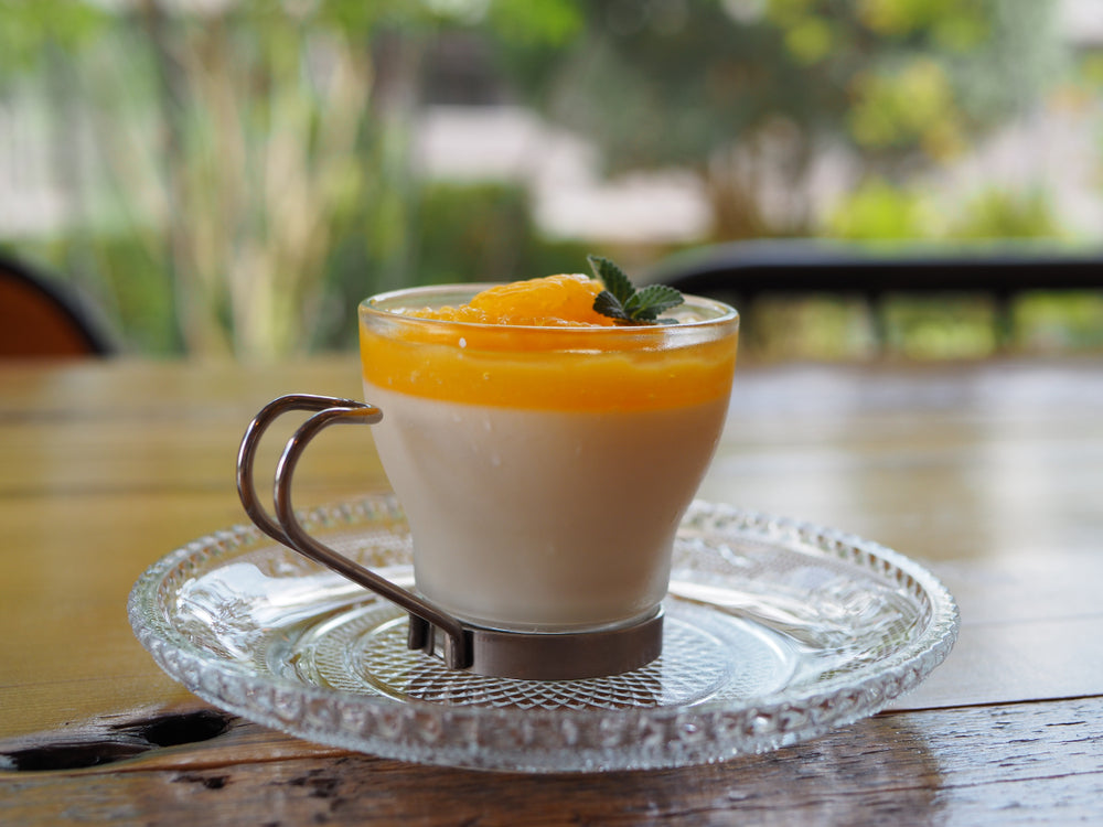 A creamy peach parfait in a glass cup, topped with orange puree and a mint leaf, sits on a decorative plate against a blurred outdoor background of greenery.