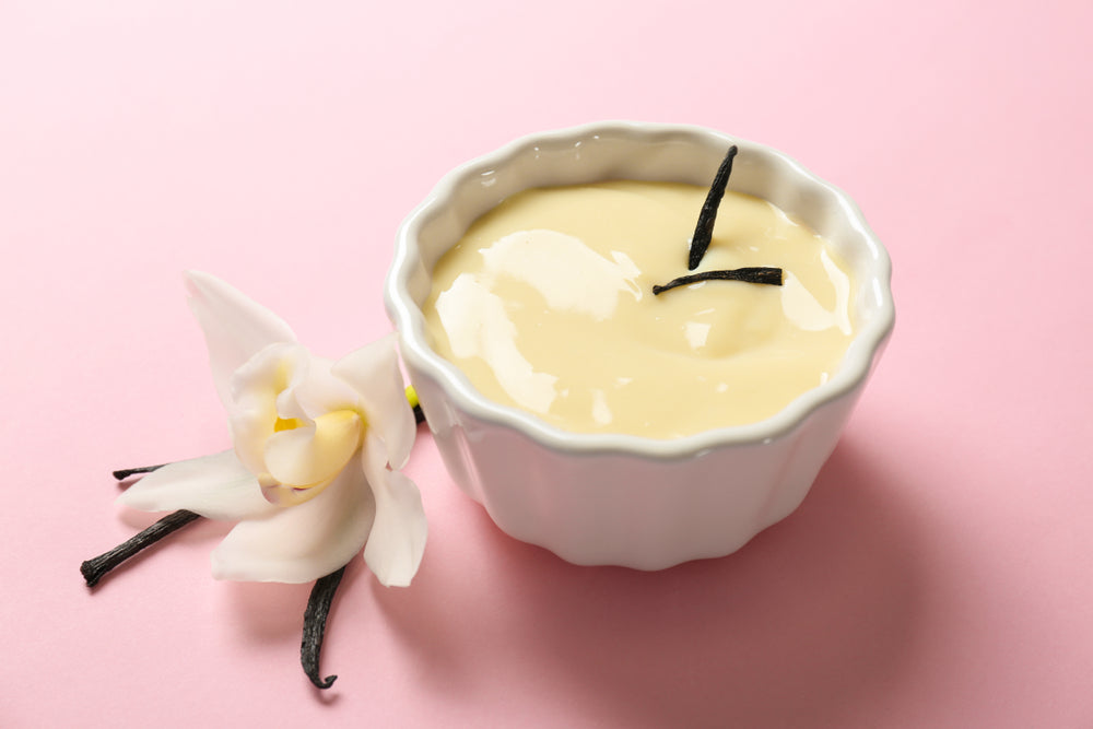 A small white bowl filled with creamy custard is placed on a pink background, accompanied by a delicate white orchid and two vanilla bean pods.