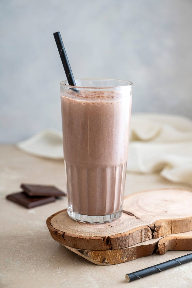A tall glass filled with creamy chocolate fudge shake sits on a wooden coaster, accompanied by chocolate squares, with a black straw resting inside the drink against a soft, neutral background.