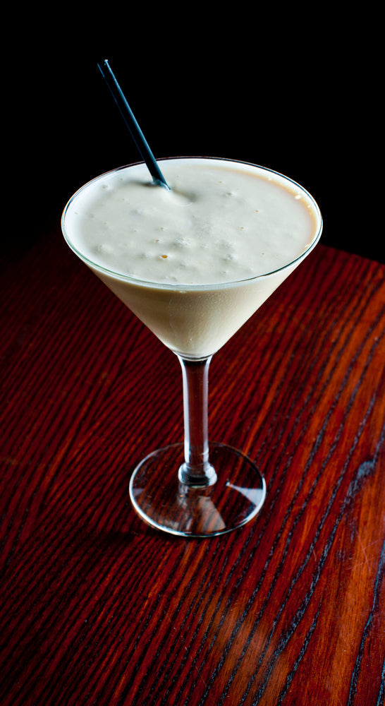 A creamy Dreamsicle Cooler cocktail sits in a clear glass on a wooden table, topped with foam and a black straw, against a dark background.
