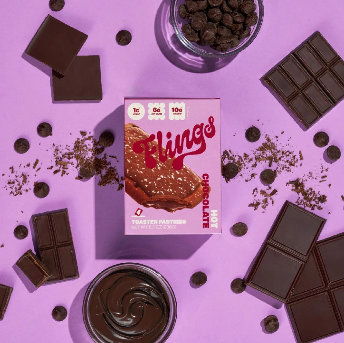 A colorful arrangement features a box of Flings Hot Chocolate Toaster Pastries surrounded by chocolate squares, chocolate chips, and a bowl of chocolate spread on a purple background.