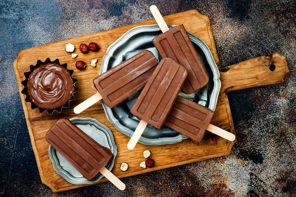 Five chocolate fudgesicles with wooden sticks are arranged on a wooden cutting board, accompanied by a bowl of chocolate sauce and scattered hazelnuts, set against a textured dark background.