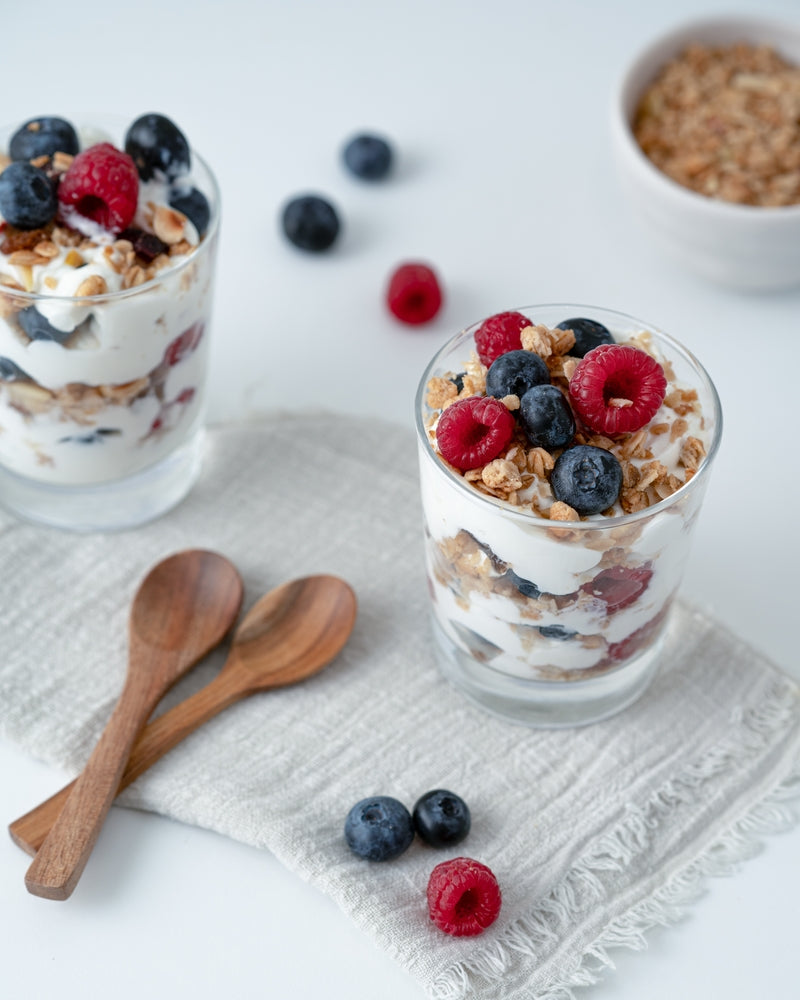 Two clear glasses filled with layers of Greek yogurt, granola, and mixed berries sit on a light fabric, accompanied by two wooden spoons and scattered blueberries and raspberries on the surface.
