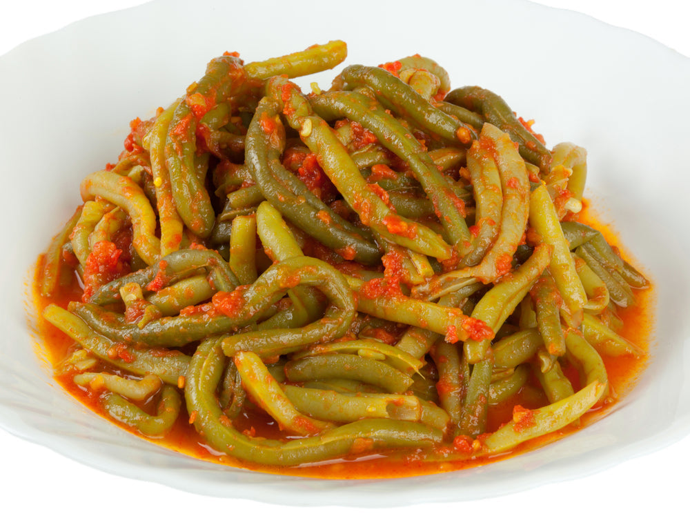 A white bowl filled with cooked green beans, coated in a rich red tomato sauce, showcases a traditional Greek dish, highlighting vibrant colors and a savory presentation.