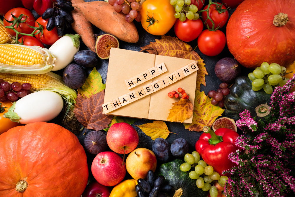 A variety of colorful fruits and vegetables surround a card with the words "Happy Thanksgiving" spelled out in wooden letters, creating a festive and healthy Thanksgiving theme.