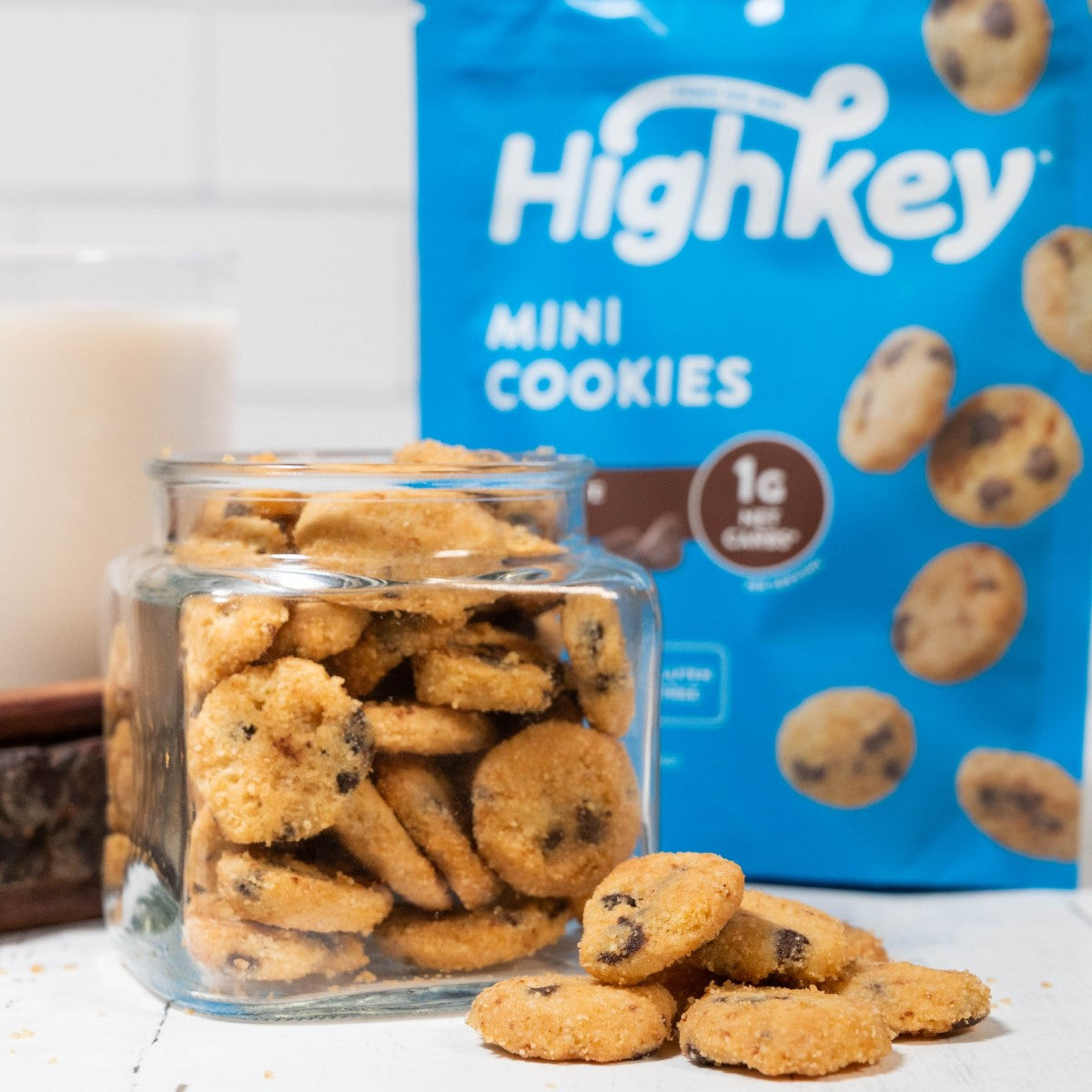 A clear glass jar filled with round mini cookies sits on a white surface, with a blue bag labeled "HighKey Mini Cookies" and a glass of milk in the background.
