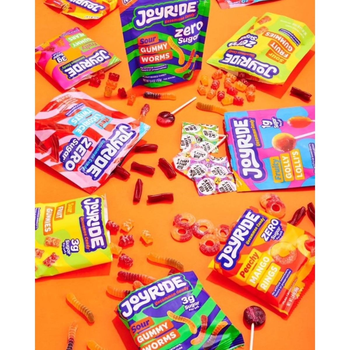 Colorful packages of Joyride candy, including gummy worms and lollipops, are scattered on a bright orange background, showcasing various flavors and sugar-free options.