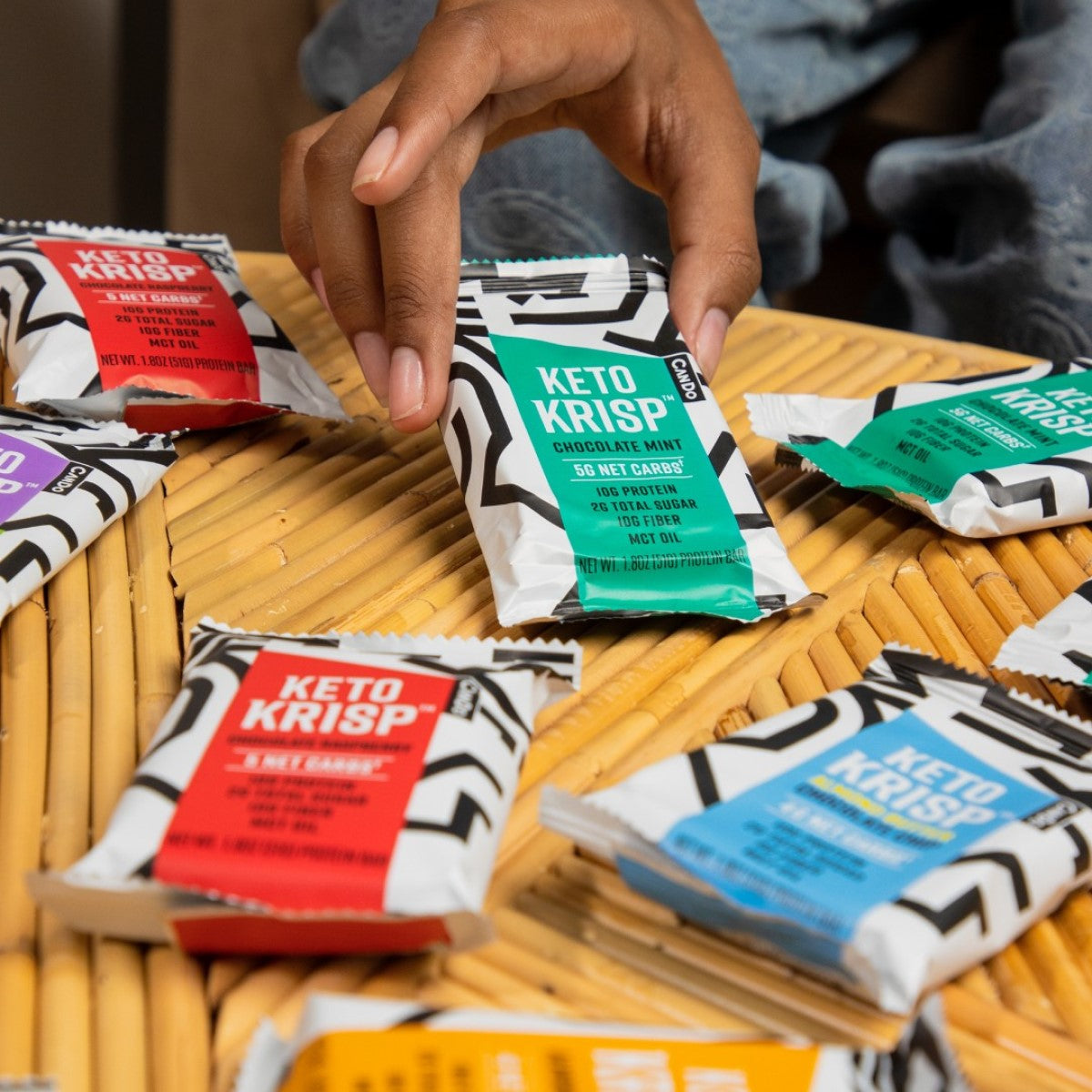 A hand reaches for a green and white Keto Krisp bar among various colorful snack wrappers on a bamboo table, showcasing a selection of low-carb protein bars.