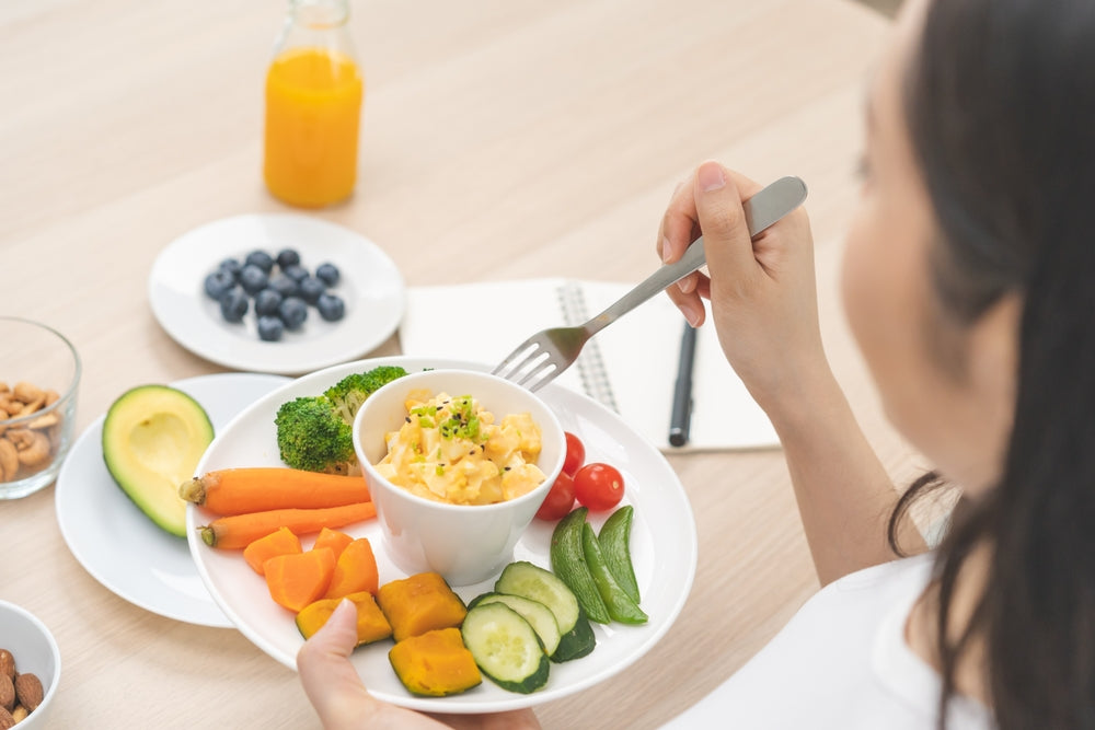 A woman holds a plate of colorful vegetables and a small bowl of food while preparing to eat, with a notebook and a glass of orange juice visible on a wooden table.