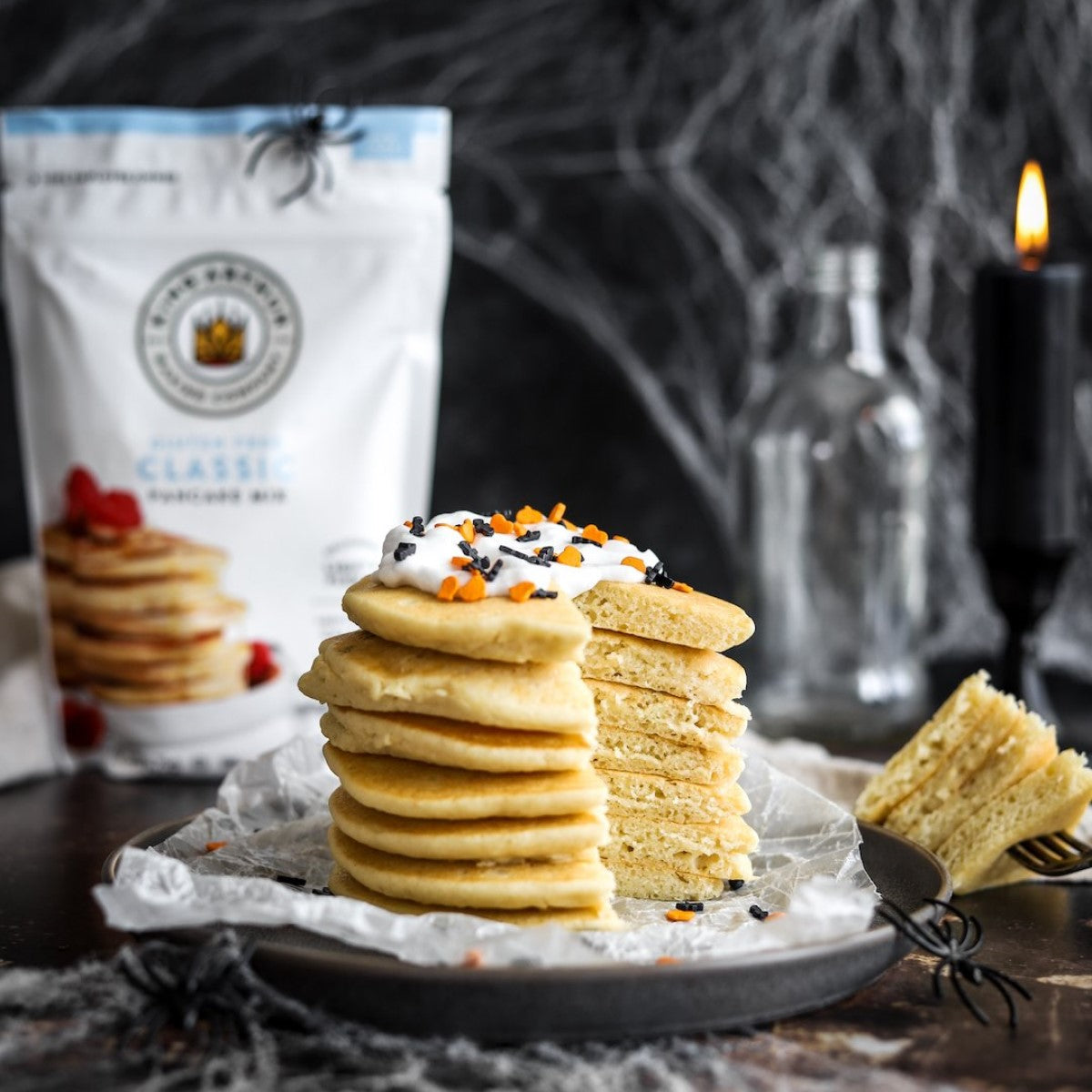 A stack of fluffy pancakes topped with whipped cream and festive sprinkles sits on a dark plate, with a bag of King Arthur Baking Company pancake mix and a candle in the background, creating a cozy atmosphere.