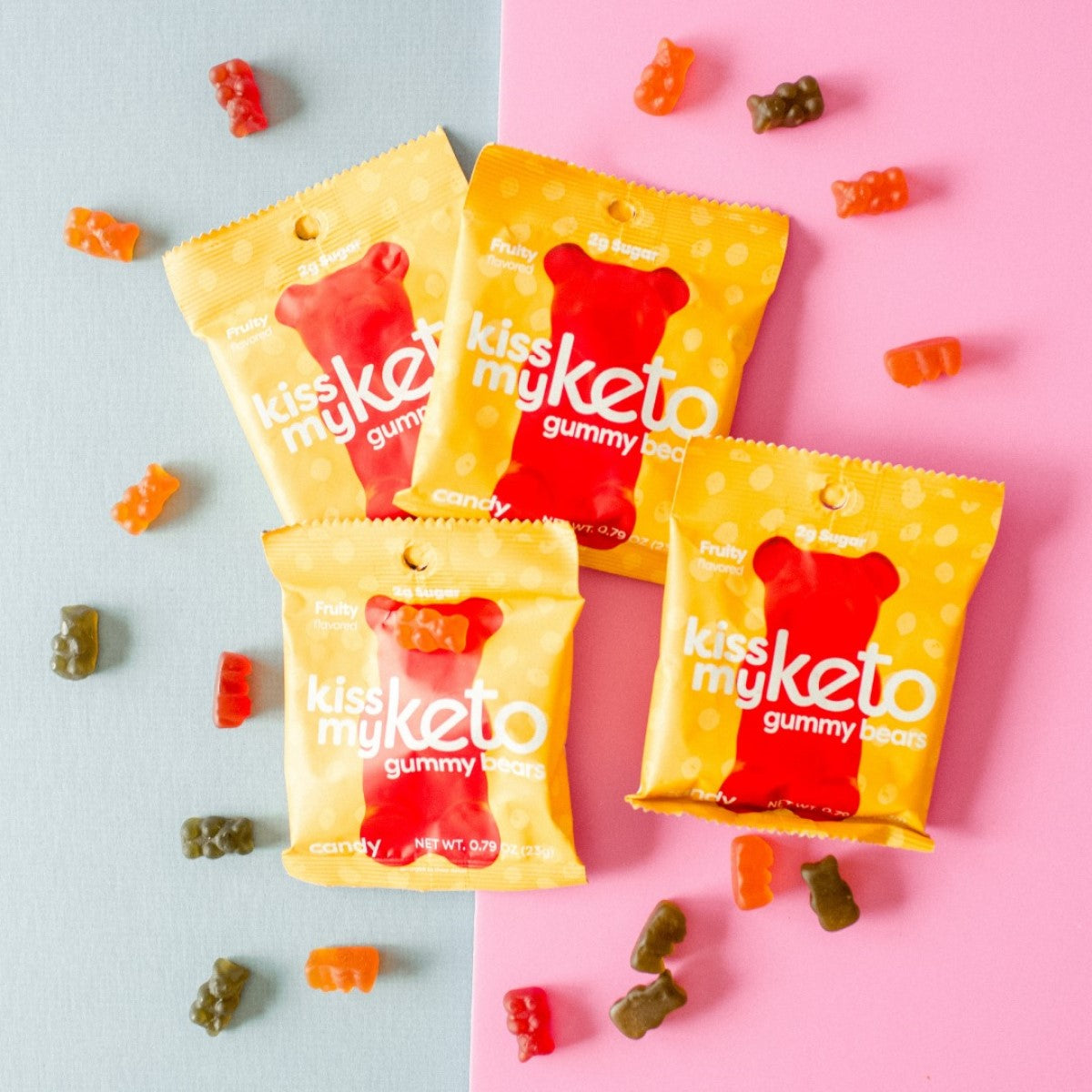 Four colorful packages of "Kiss My Keto" gummy bears are arranged on a light blue and pink background, with several gummy bears scattered around, showcasing their fruity flavors.