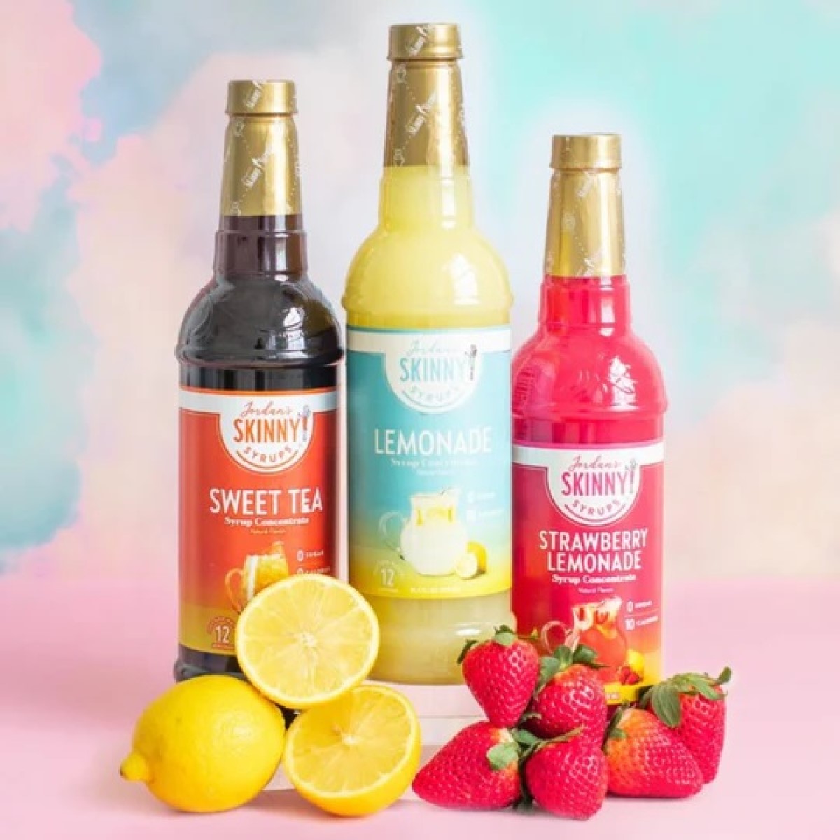 Three bottles of Jordan's Skinny Syrups—Sweet Tea, Lemonade, and Strawberry Lemonade—are displayed on a pastel pink surface, accompanied by fresh lemons and strawberries, against a soft, colorful background.