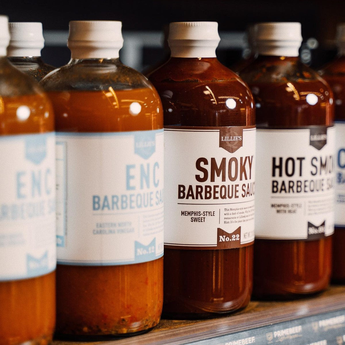 Several bottles of Lillie's Q Zero Sugar BBQ sauces are displayed on a wooden shelf, featuring labels that highlight different flavors like Smoky and Hot Smoky, emphasizing their Memphis-style recipes.