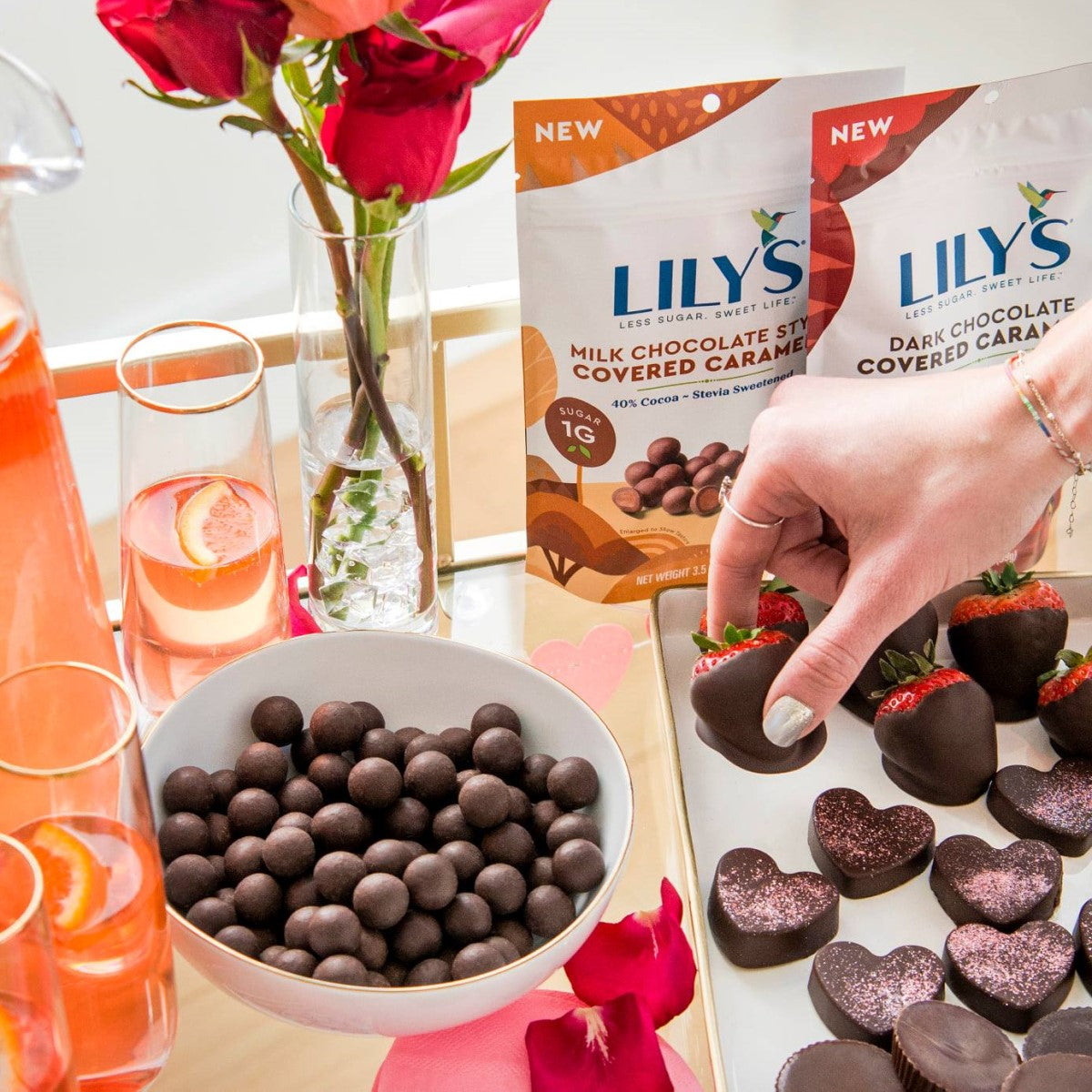 A hand reaches for a chocolate-covered strawberry from a tray, surrounded by bowls of chocolate candies, heart-shaped chocolates, and two bags of Lily's Sweets, with pink flowers and drinks in the background.