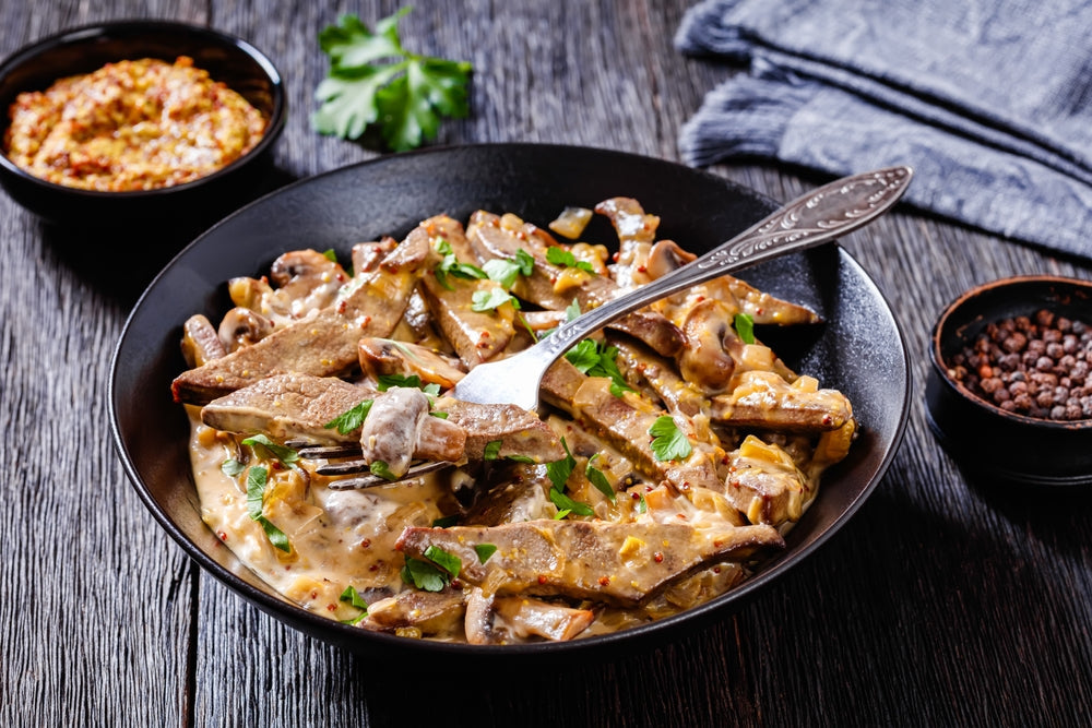 A black bowl filled with liver and onions in a creamy sauce, garnished with parsley, sits on a wooden table alongside small bowls of mustard and spices.