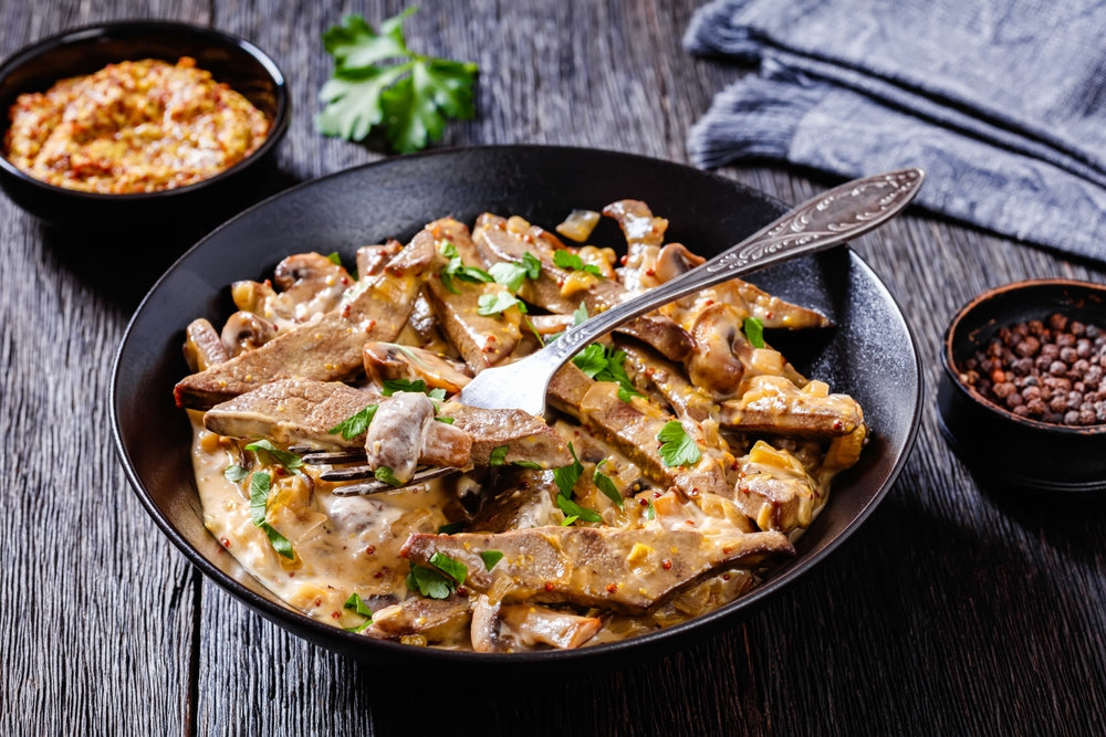 A black bowl filled with liver and onions in a creamy sauce, garnished with parsley, sits on a wooden table alongside small bowls of mustard and spices.