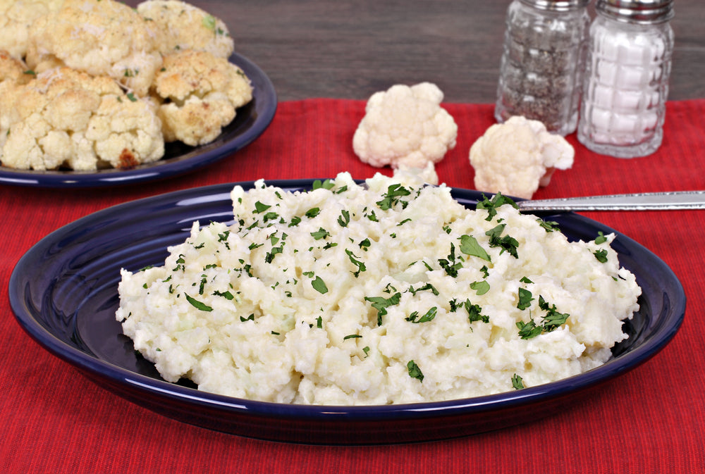 A blue plate holds a serving of mock mashed potatoes, garnished with green herbs, next to a plate of roasted cauliflower, with salt shakers in the background on a red tablecloth.