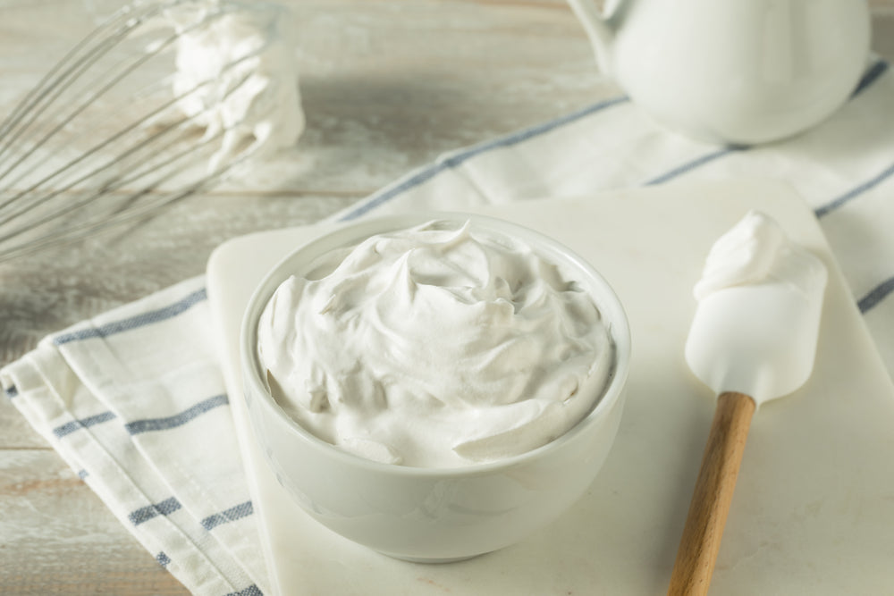 A bowl of fluffy, white mock whipped topping sits on a cutting board, accompanied by a spatula and a whisk, with a striped towel in the background on a rustic wooden surface.
