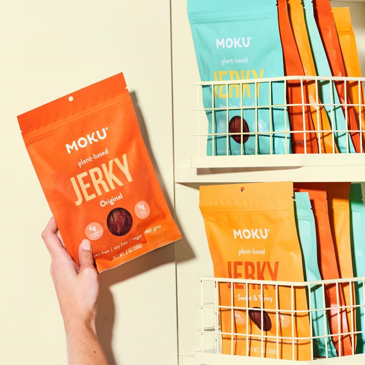 A hand holds a bright orange package of Moku plant-based jerky labeled "Original," while colorful bags of jerky in various flavors are neatly arranged on a white shelf behind it.