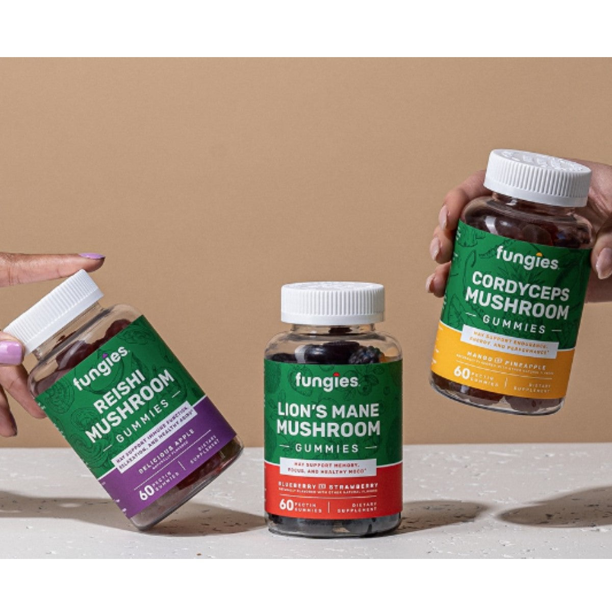 Three bottles of Fungies mushroom gummies are displayed on a light surface; one hand holds a Reishi bottle, while another hand presents a Cordyceps bottle, with a Lion's Mane bottle in between.