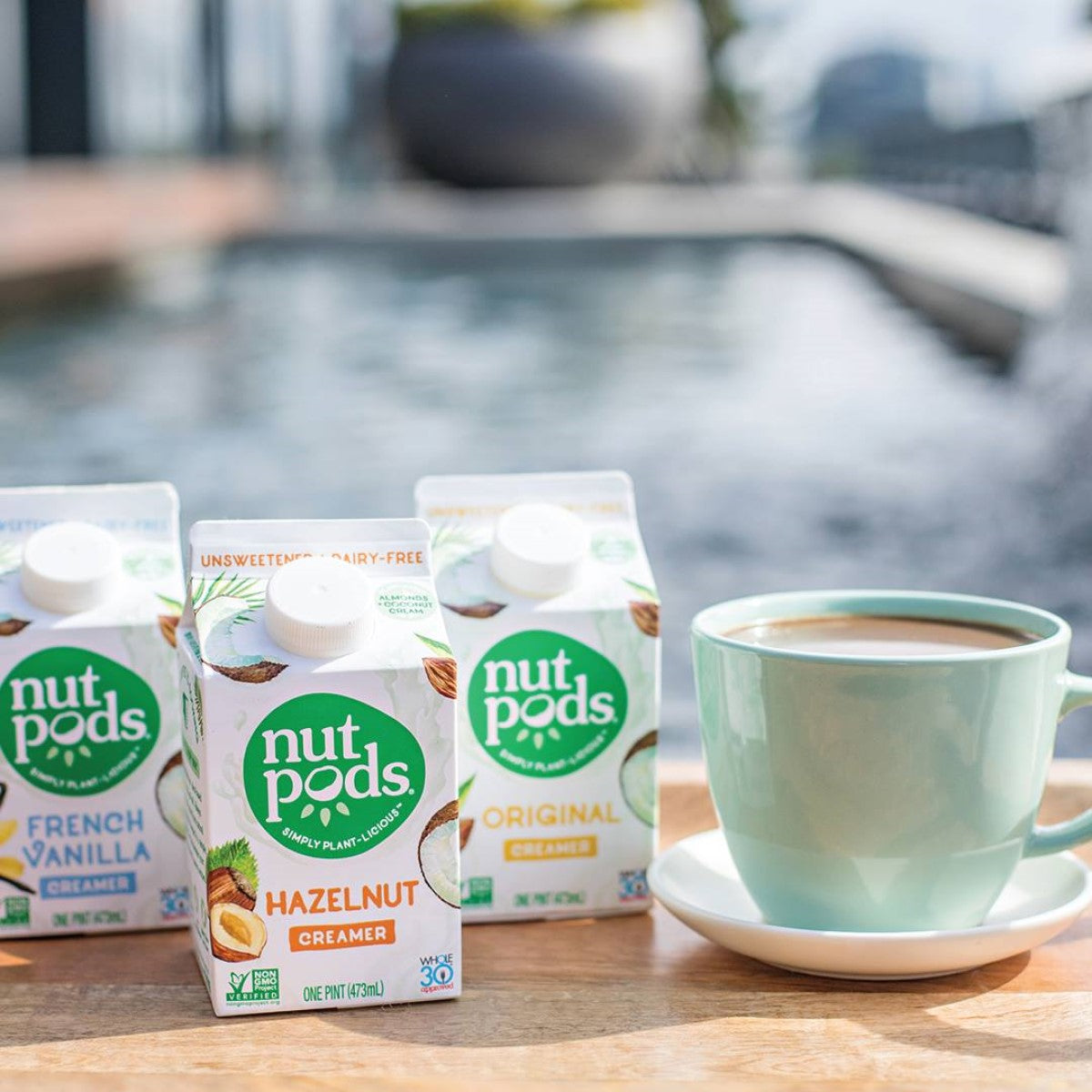 Three cartons of Nutpods dairy-free creamers (Hazelnut, French Vanilla, Original) are displayed on a wooden tray beside a light green cup of coffee, with a blurred water feature and greenery in the background.