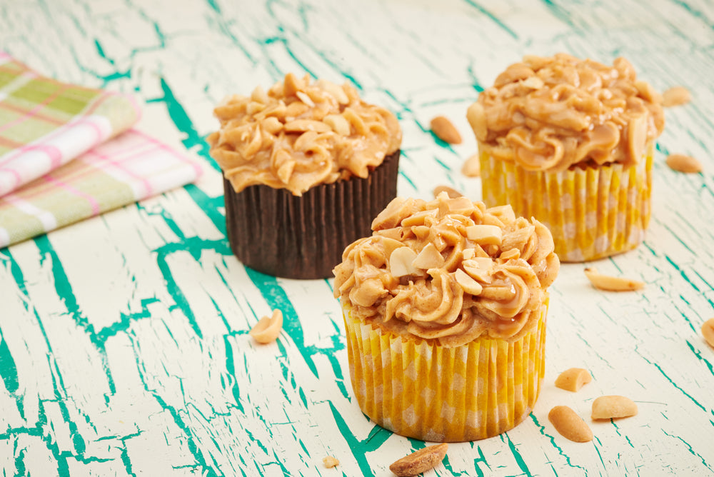 Three peanut butter cupcakes topped with creamy frosting and chopped peanuts sit on a rustic, teal-painted wooden surface, with a checkered napkin nearby and scattered peanuts around.