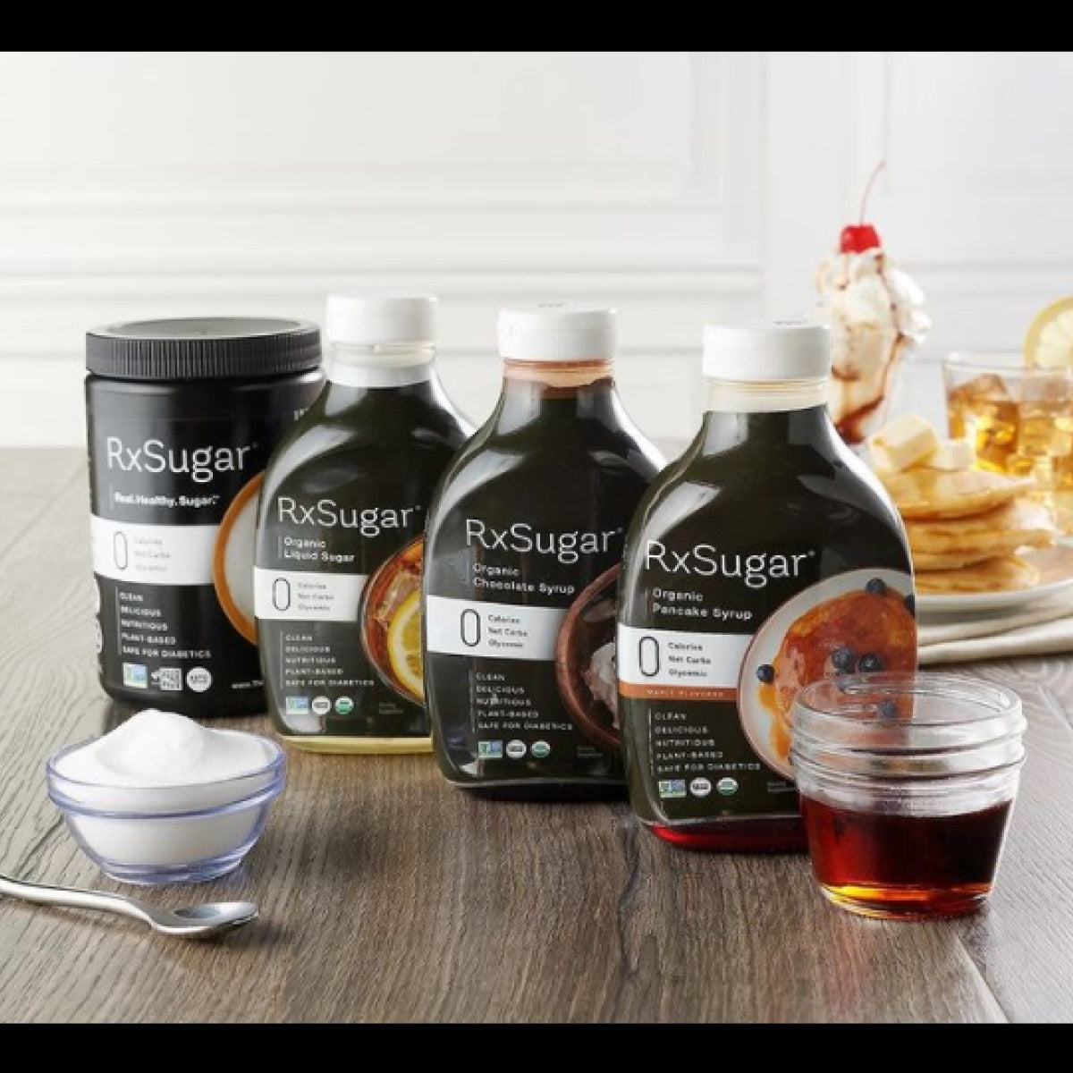 Four bottles of RxSugar organic sweeteners and syrups are displayed on a wooden table, alongside a small bowl of sugar and a glass of syrup, with pancakes in the background.