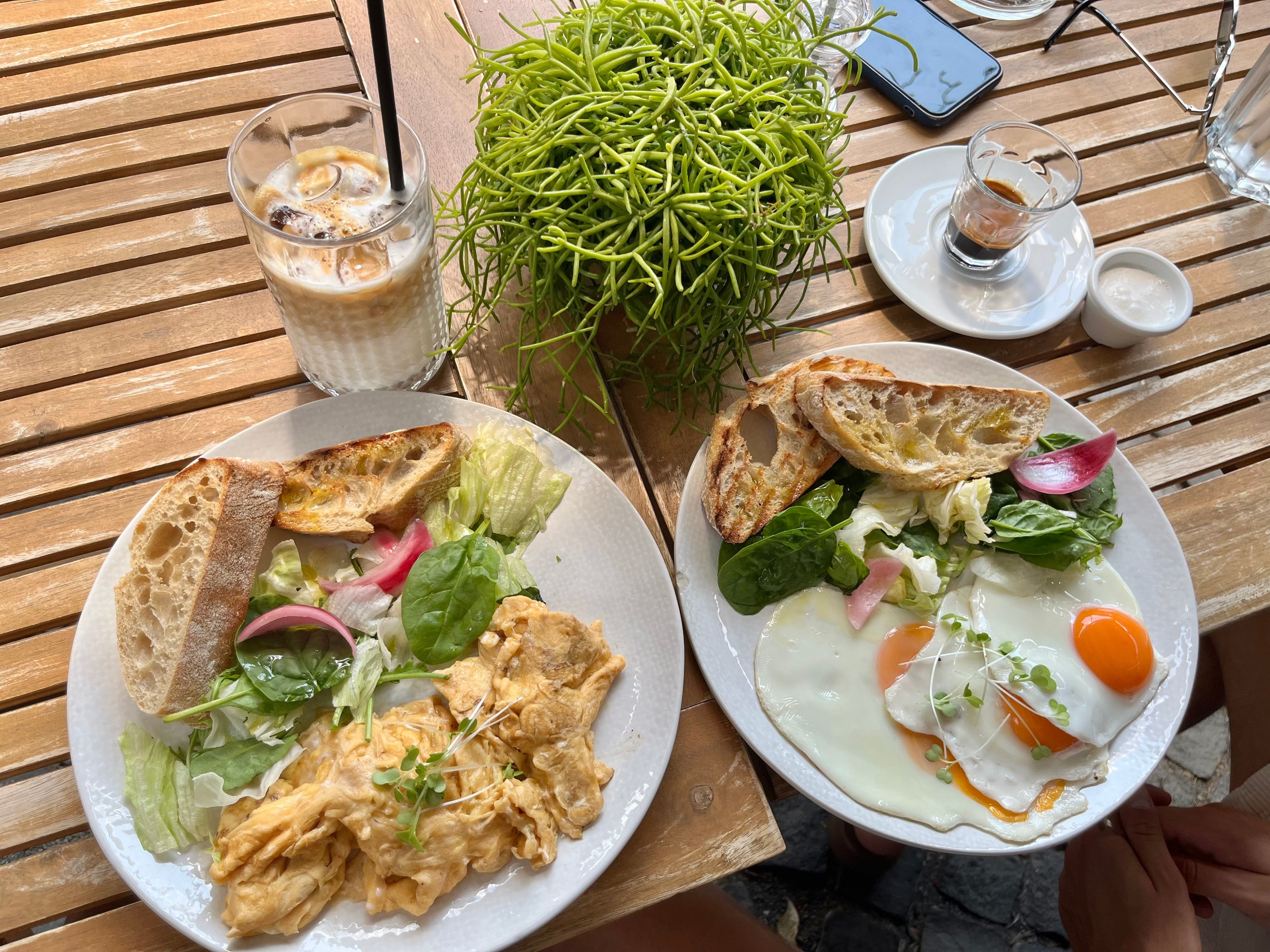 Two plates of breakfast food featuring scrambled eggs, fried eggs, salad, and toasted bread are set on a wooden table, accompanied by a glass of iced coffee and a small dish of cream.