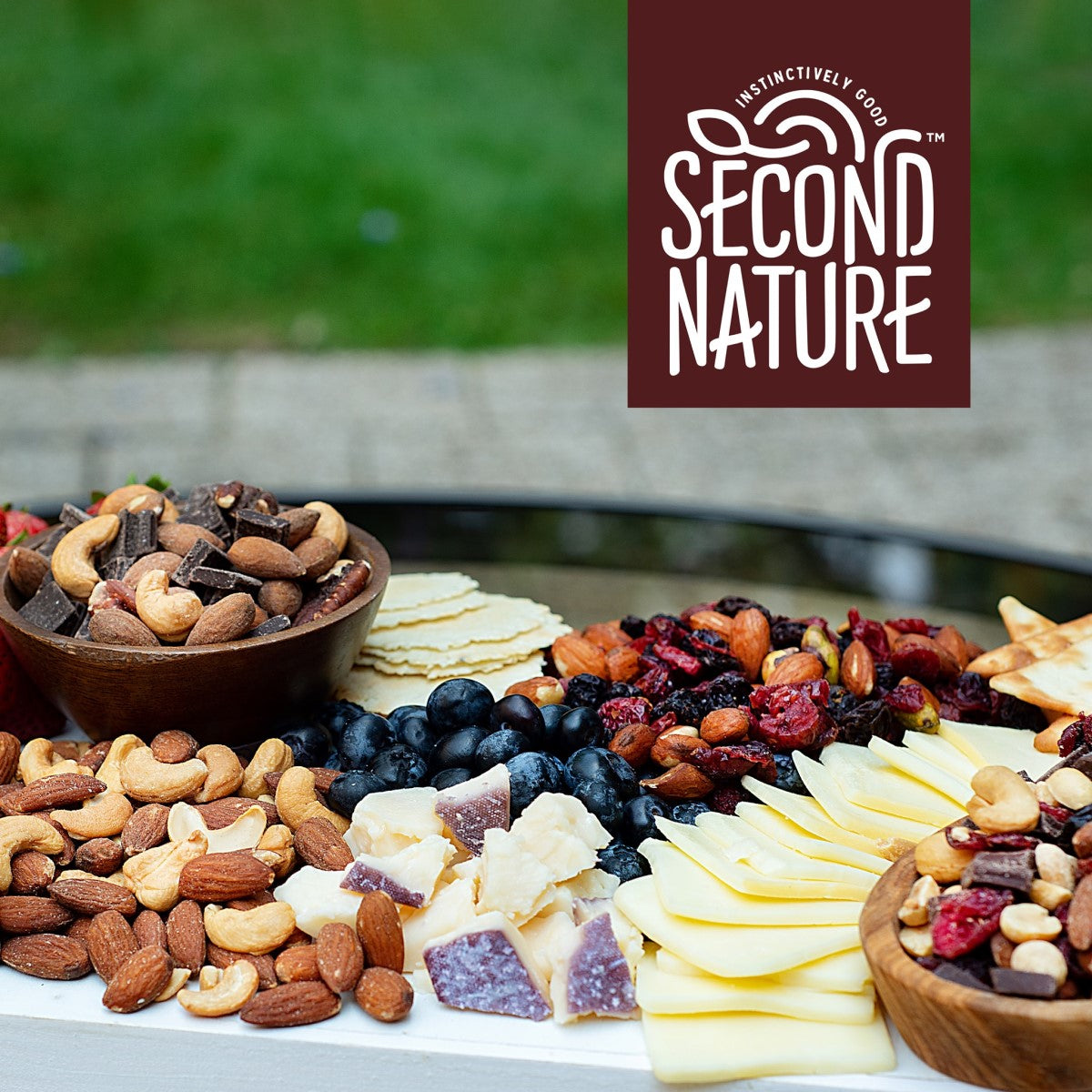 A colorful assortment of nuts, cheeses, dried fruits, and crackers is displayed on a wooden platter outdoors, with the Second Nature logo prominently featured in the corner.