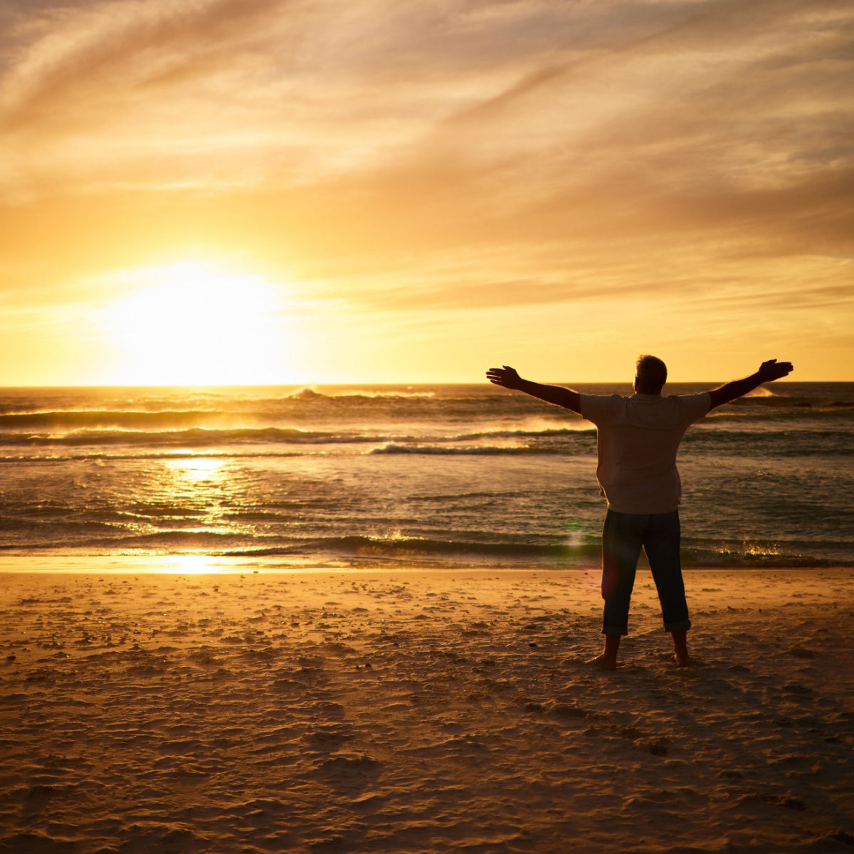 A person stands barefoot on a sandy beach at sunset, arms outstretched, gazing at the ocean waves, symbolizing freedom and new beginnings after bariatric surgery.