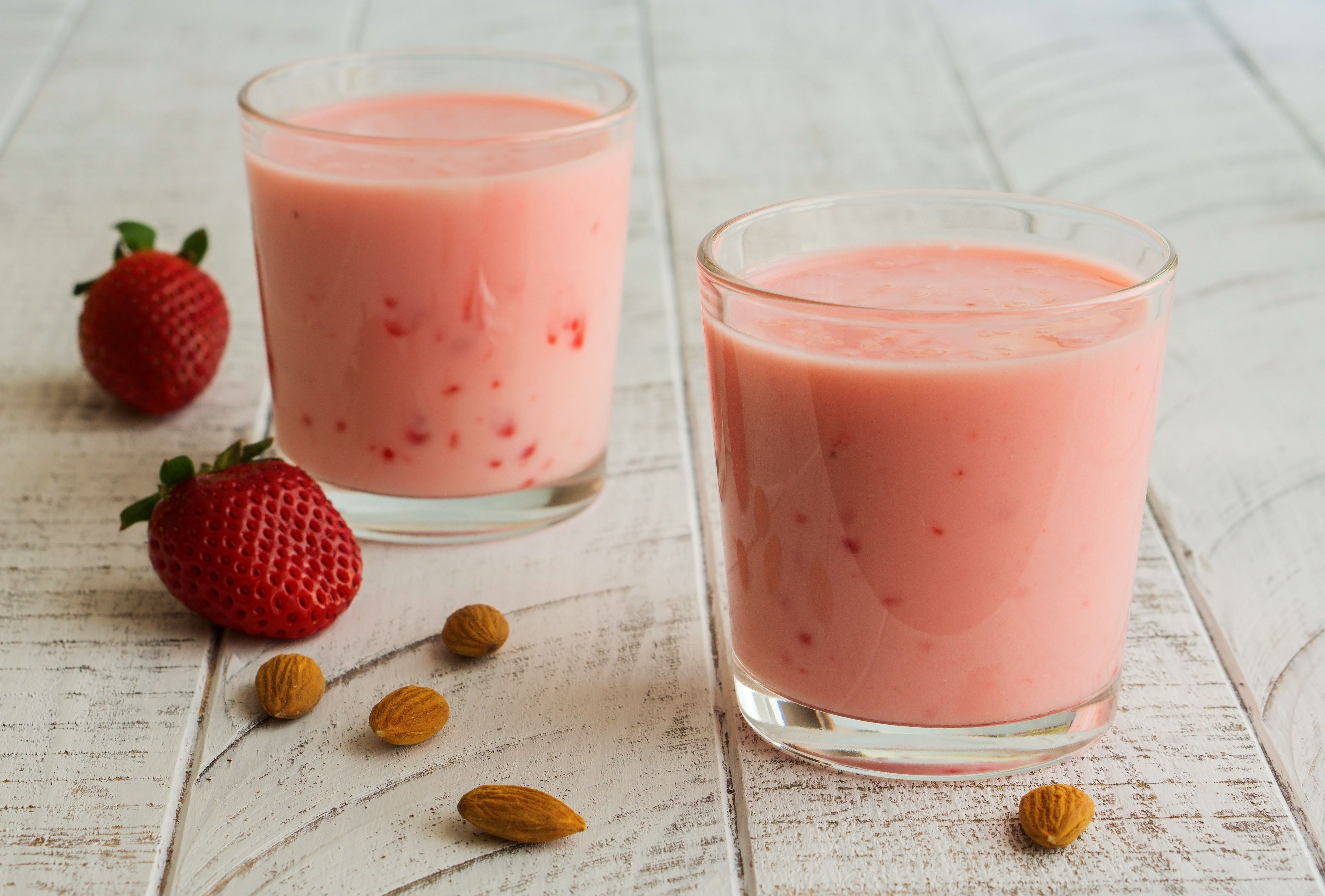 Two glasses of pink smoothie sit on a white wooden surface, surrounded by fresh strawberries and almonds, suggesting a healthy, fruity drink option.