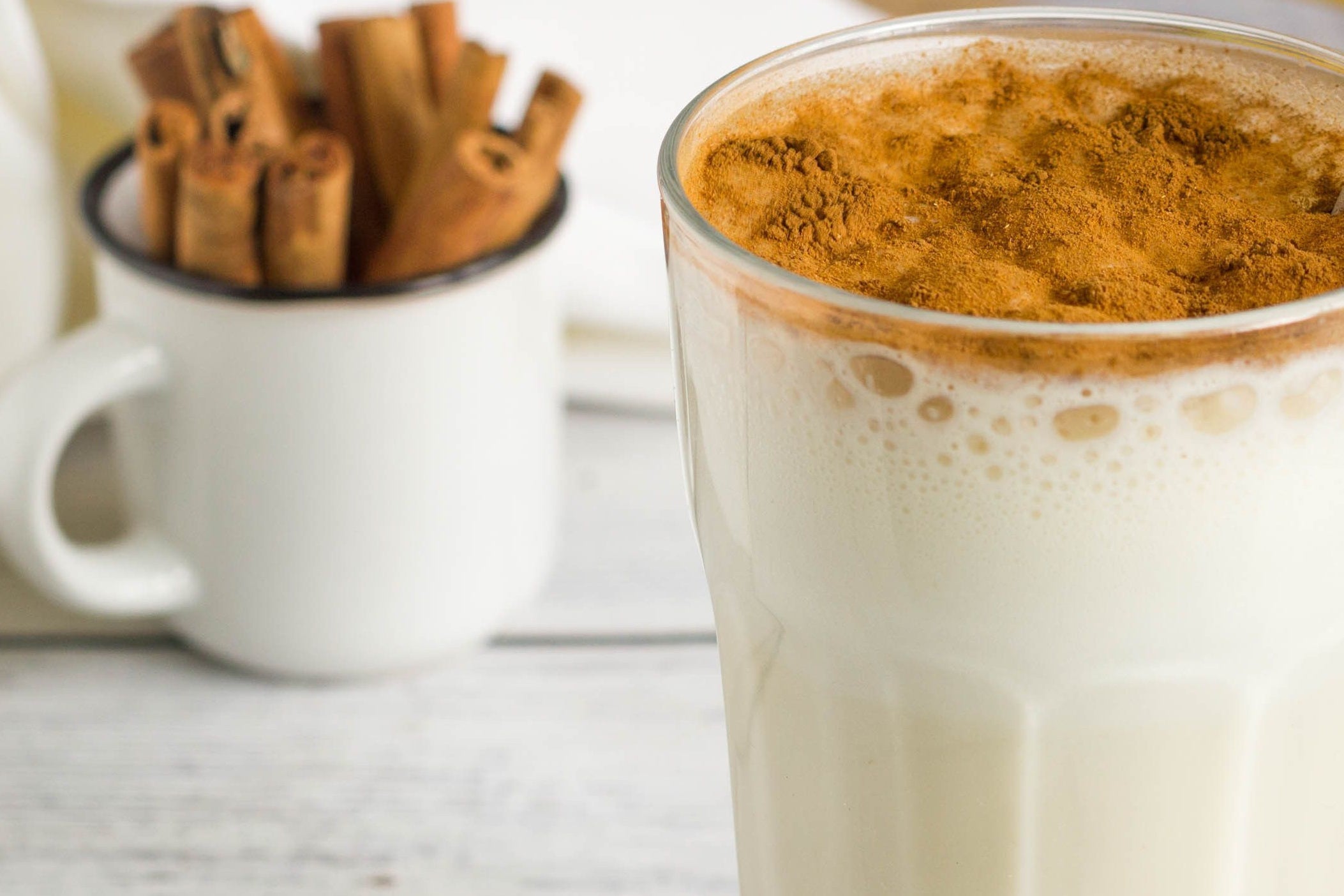 A glass of creamy maple smoothie topped with a sprinkle of cinnamon sits on a wooden surface, with a cup of cinnamon sticks in the background.