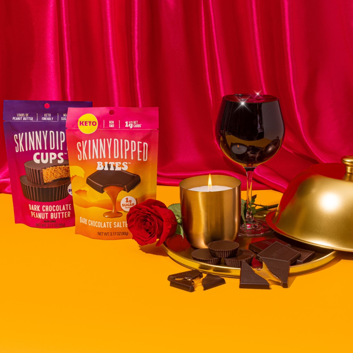Two packages of SkinnyDipped chocolate products are displayed alongside a glass of red wine, a lit candle, a gold serving dish, and a red rose, set against a vibrant orange background and pink satin curtain.