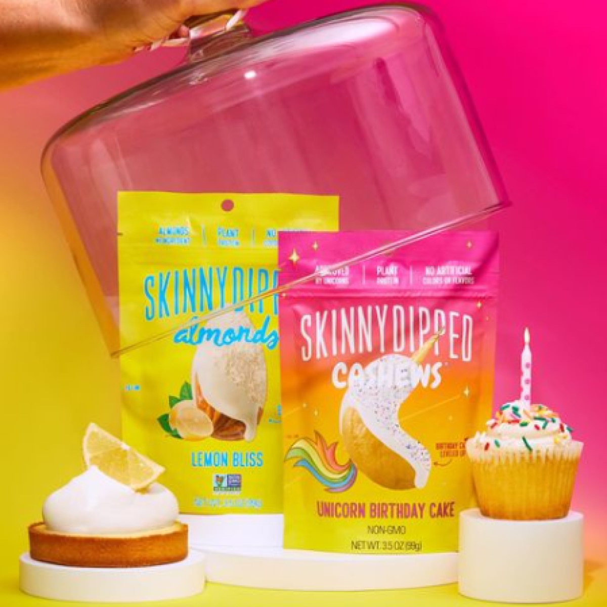 A hand lifts a glass dome revealing colorful packages of SkinnyDipped Almonds and Cashews, alongside a cupcake with a candle and a lemon tart, set against a vibrant pink and yellow background.