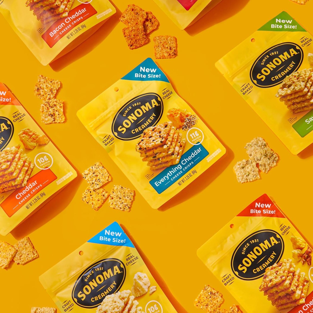Colorful packages of Sonoma Creamery cheese crisps in various flavors, surrounded by scattered crisps, are displayed against a bright yellow background, highlighting their new bite-sized format.