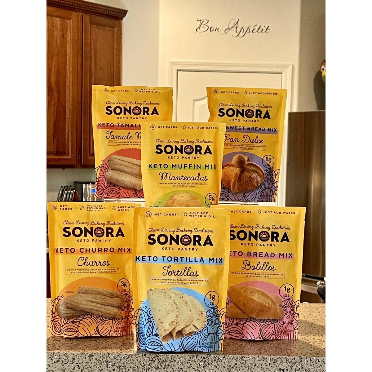 A display of various Sonora Keto Pantry baking mixes, including tamale, muffin, churro, tortilla, sweet bread, and bolillo mixes, arranged on a kitchen counter with a "Bon Appétit" sign in the background.